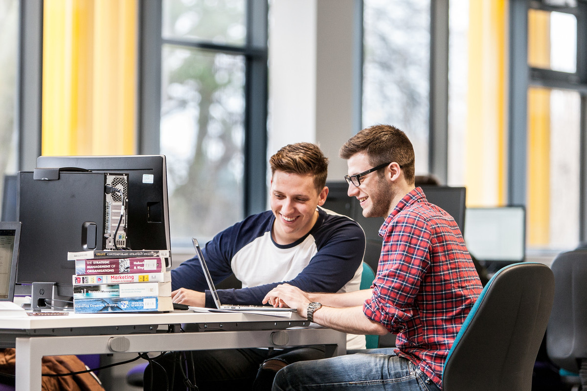 Two young male students sitting at a table with books and laptop. Both are smiling looking at the laptop screen