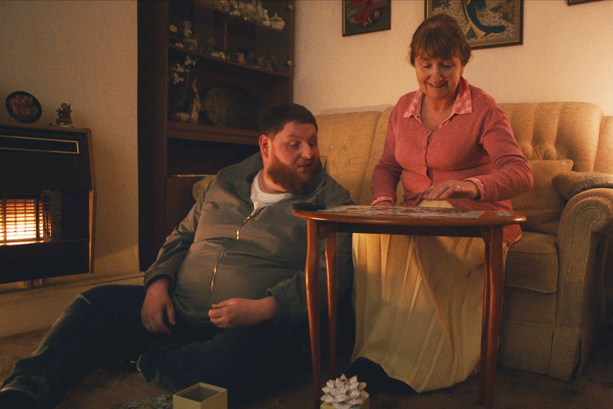 A still from a featured film, with a man sat down looking at a jigsaw being completed by a woman to his left.
